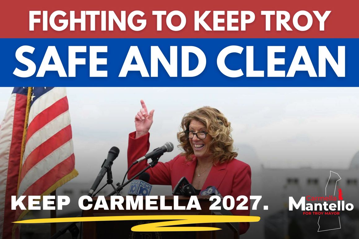 Campaign graphic reading "Fighting to keep Troy safe and clean" and "Keep Carmella 2027." Mayor Carmella Mantello speaks at a podium of news microphones beside an American flag.